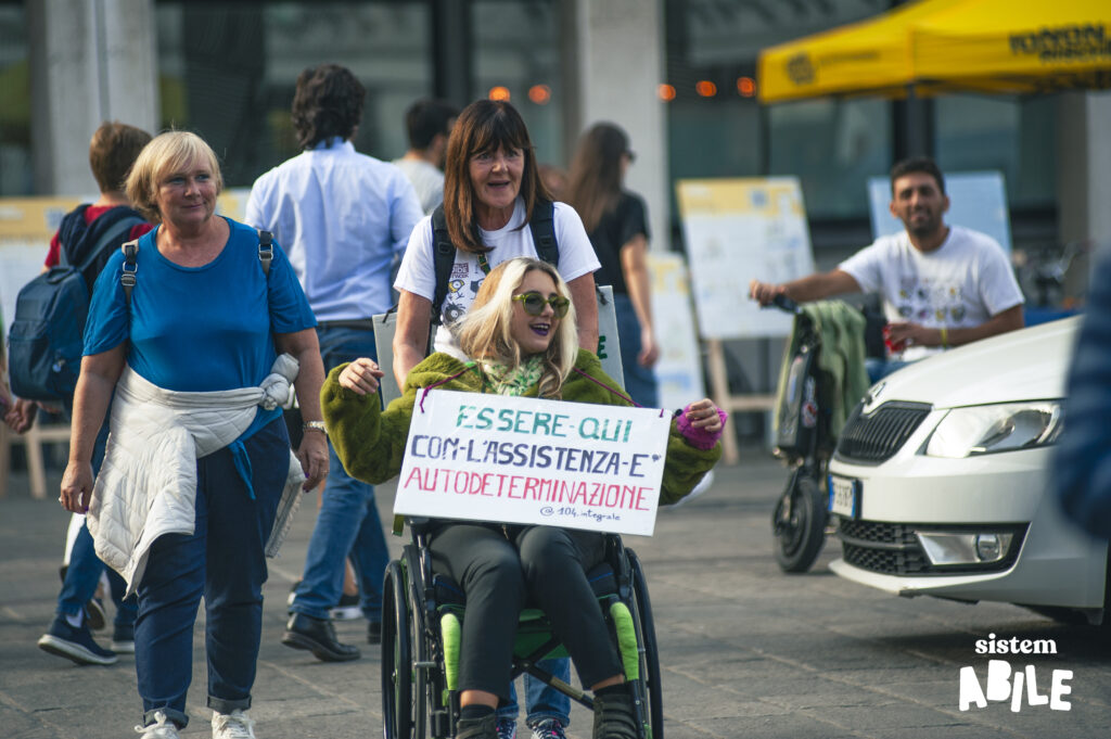 Una persona raffigurata al corteo del disability pride. Sul cartello c'è scritto: "Essere qui con l'assistenza è autodeterminazione"