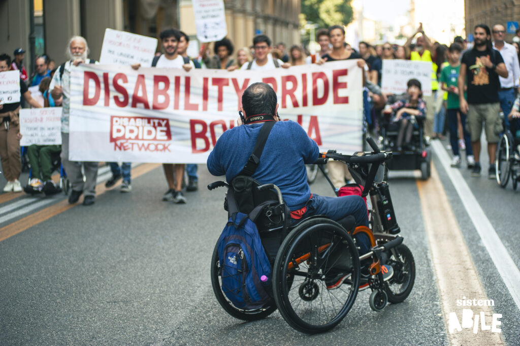 Una persona sulla sedia a rotelle fotografa lo striscione che apre il corte del disability pride